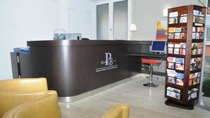 Un mostrador de recepción con un cartel que dice 'Hotel B', una computadora, una estantería con revistas y dos sillas amarillas.