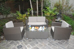 Un patio con mobiliario de mimbre, incluyendo un sofá y dos sillones, y una mesa de centro de vidrio con cosas para el desayuno.