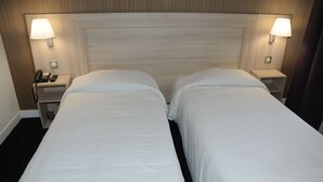 Habitación de hotel con dos camas individuales, cabecero de madera, lámparas de noche, teléfono y una mesita.