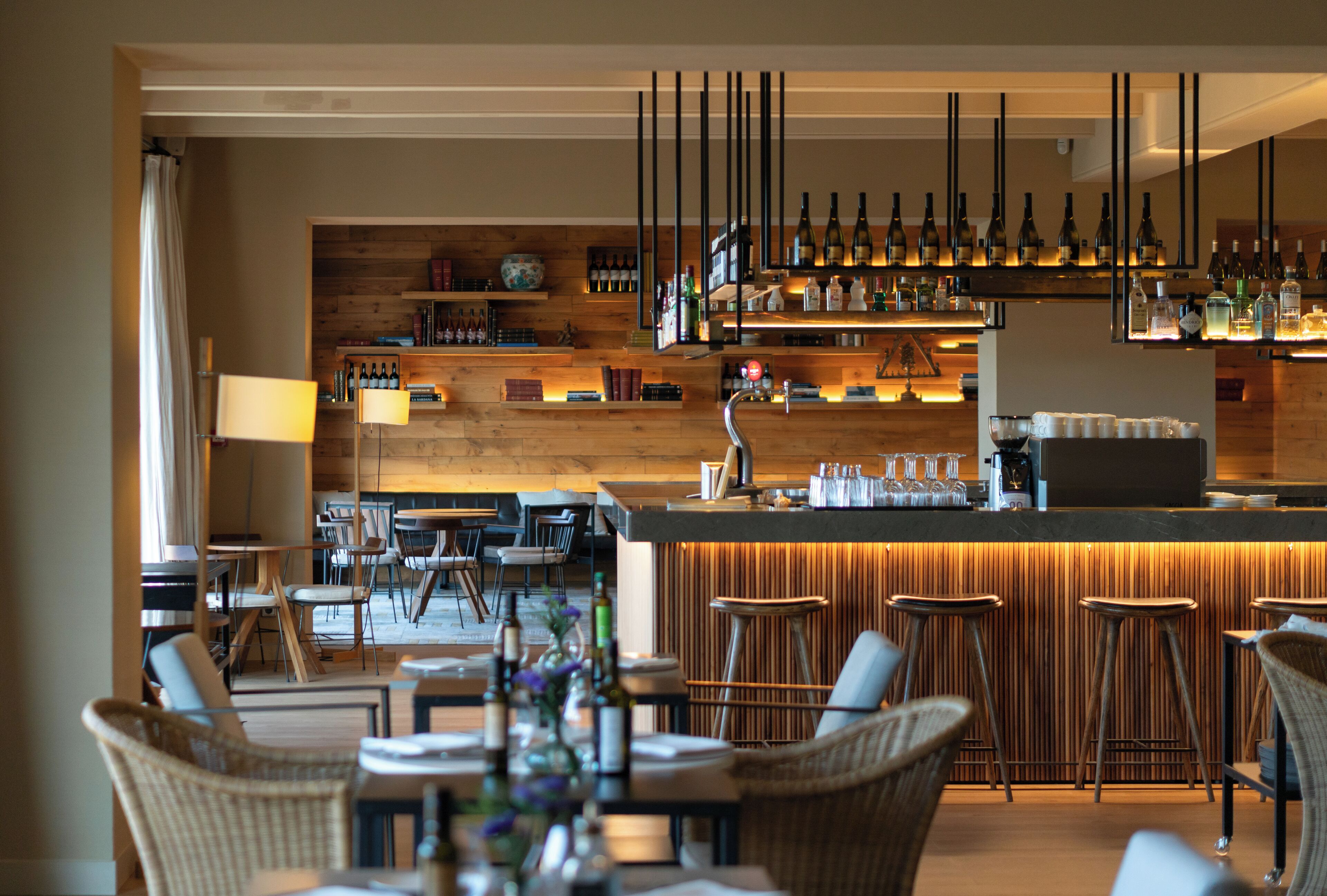 Zona de bar con estantes de madera, botellas colgantes y taburetes.