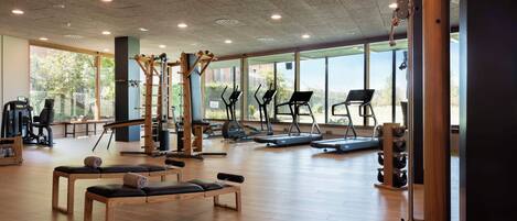A spacious gym with various exercise equipment, large windows, and a wooden floor.