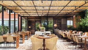 An outdoor dining area with wicker furniture, a thatched roof, and a view of greenery.