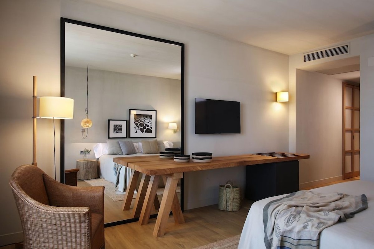 A modern hotel room with a wooden desk, a large mirror, a television, and a comfortable seating area with a sofa and a chair.