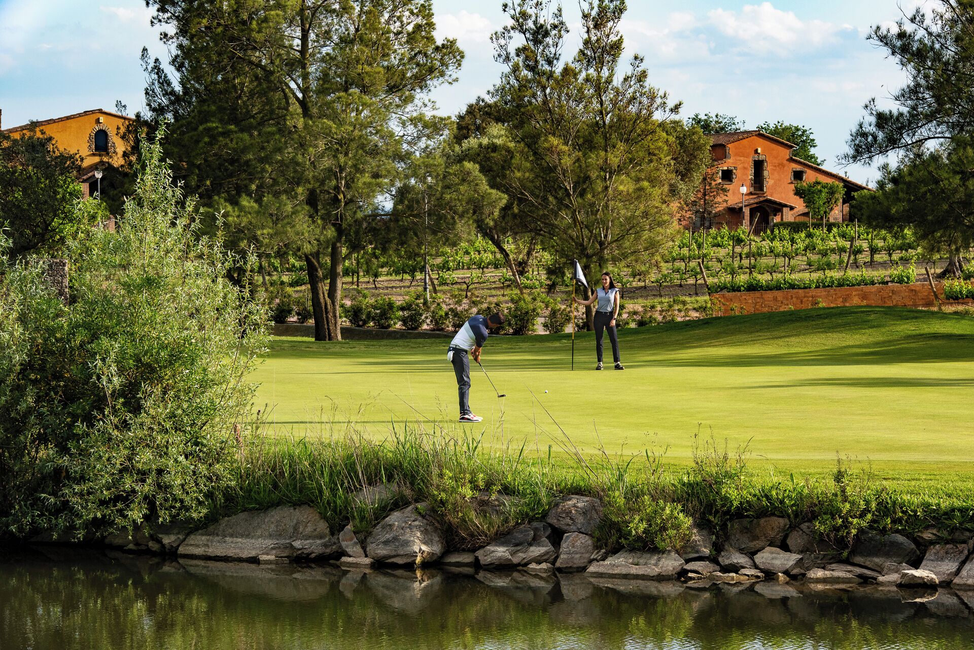 Un campo de golf con un estanque, árboles y edificios al fondo.