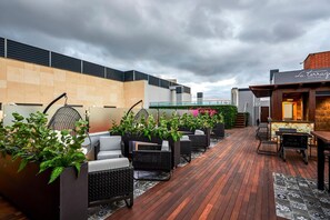 An outdoor seating area with wicker furniture and potted plants, a bar area with a neon sign, and a modern building facade. - Gran Hotel Victoria (El Ejido)
