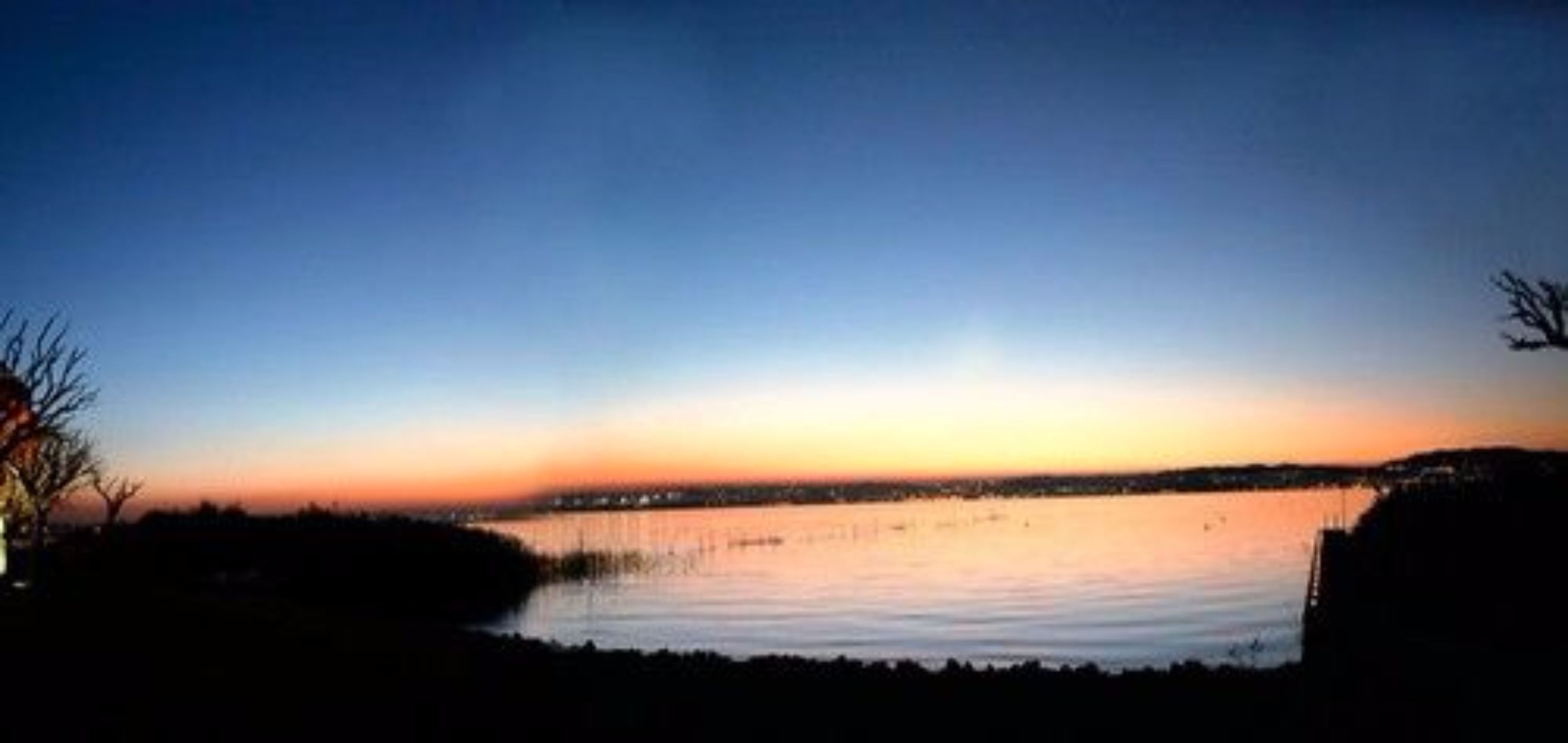 Sonnenuntergang über ruhigem Gewässer mit einer fernen Küste und der Silhouette eines Baumes.