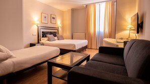 Quarto de hotel com duas camas de solteiro, um sofá, uma mesa de centro de vidro, uma televisão e um radiador.