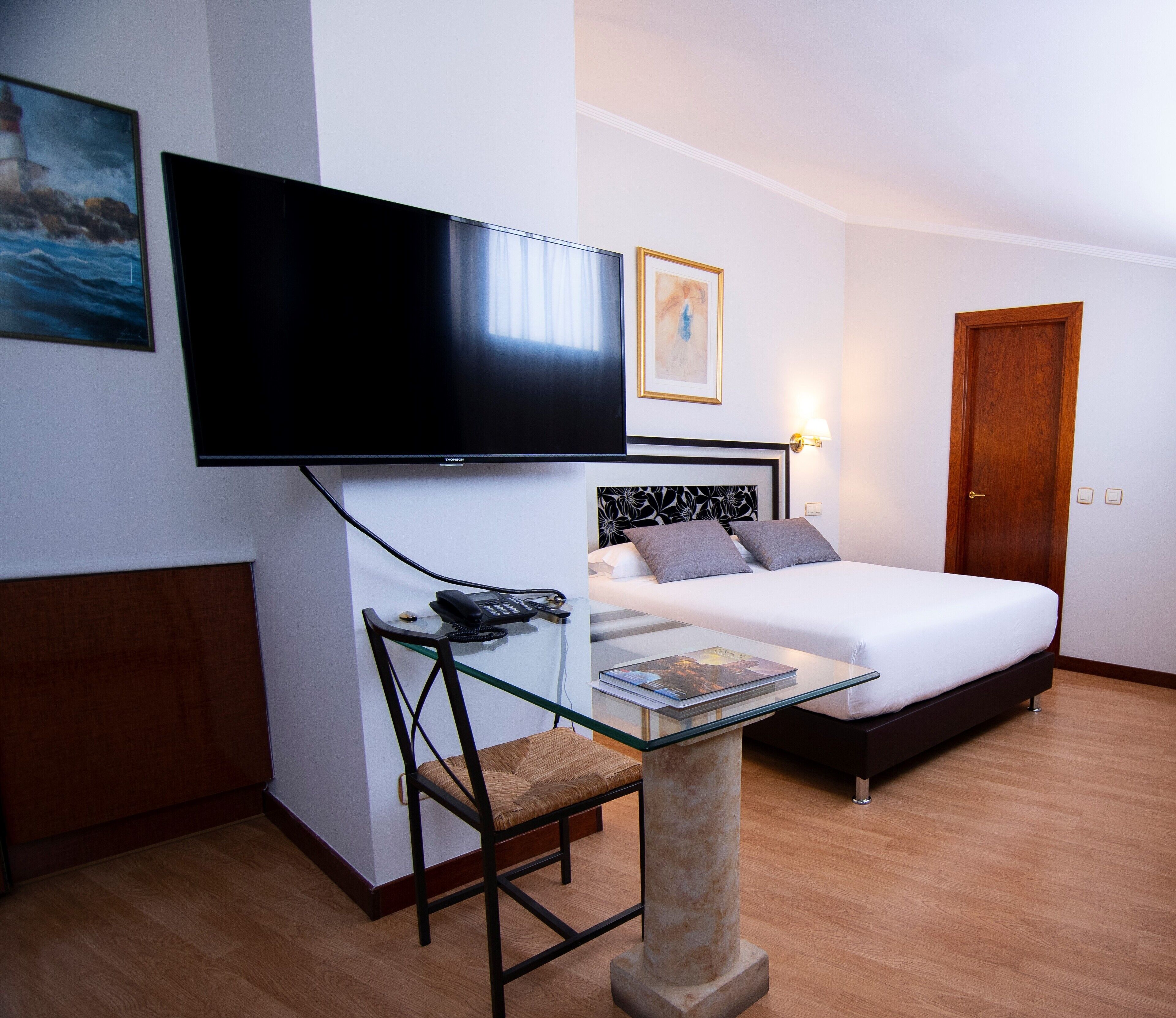 A hotel room with a bed, a desk with a chair, a television mounted on the wall, and a door.