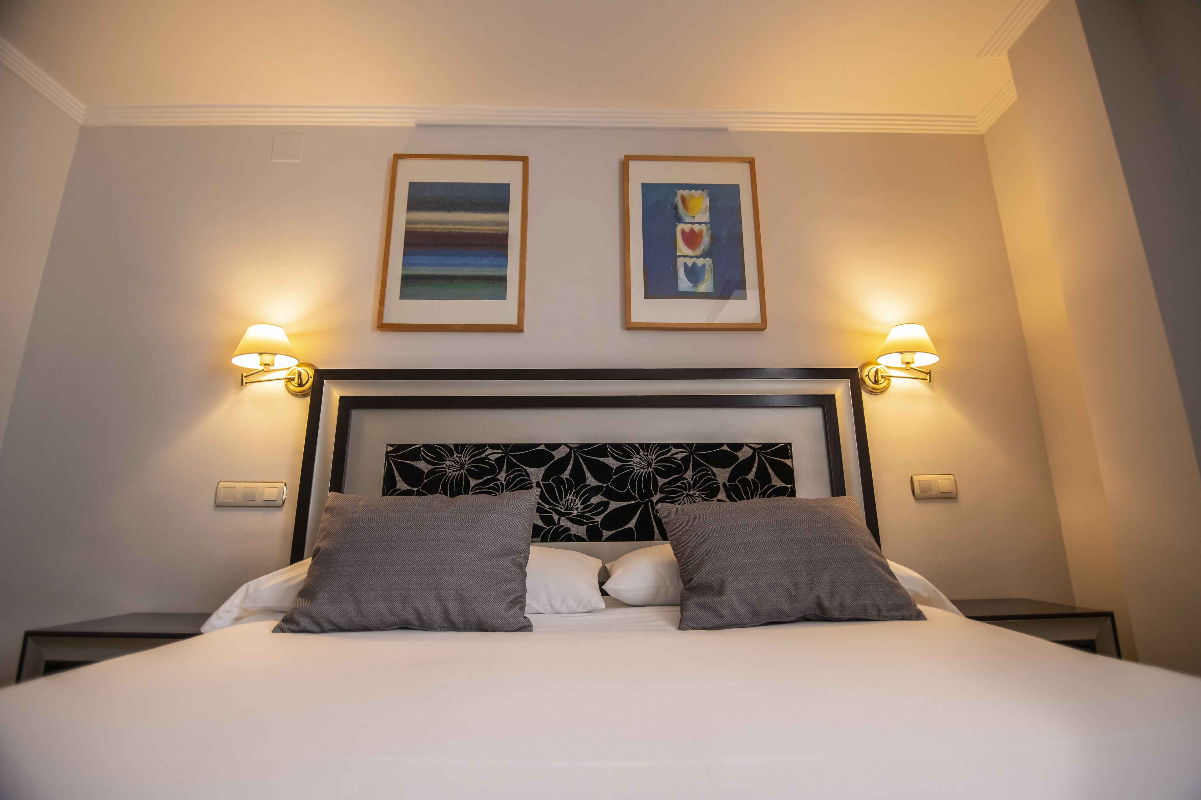 A neatly made bed with two pillows, a headboard with a floral pattern, and two framed pictures on the wall.