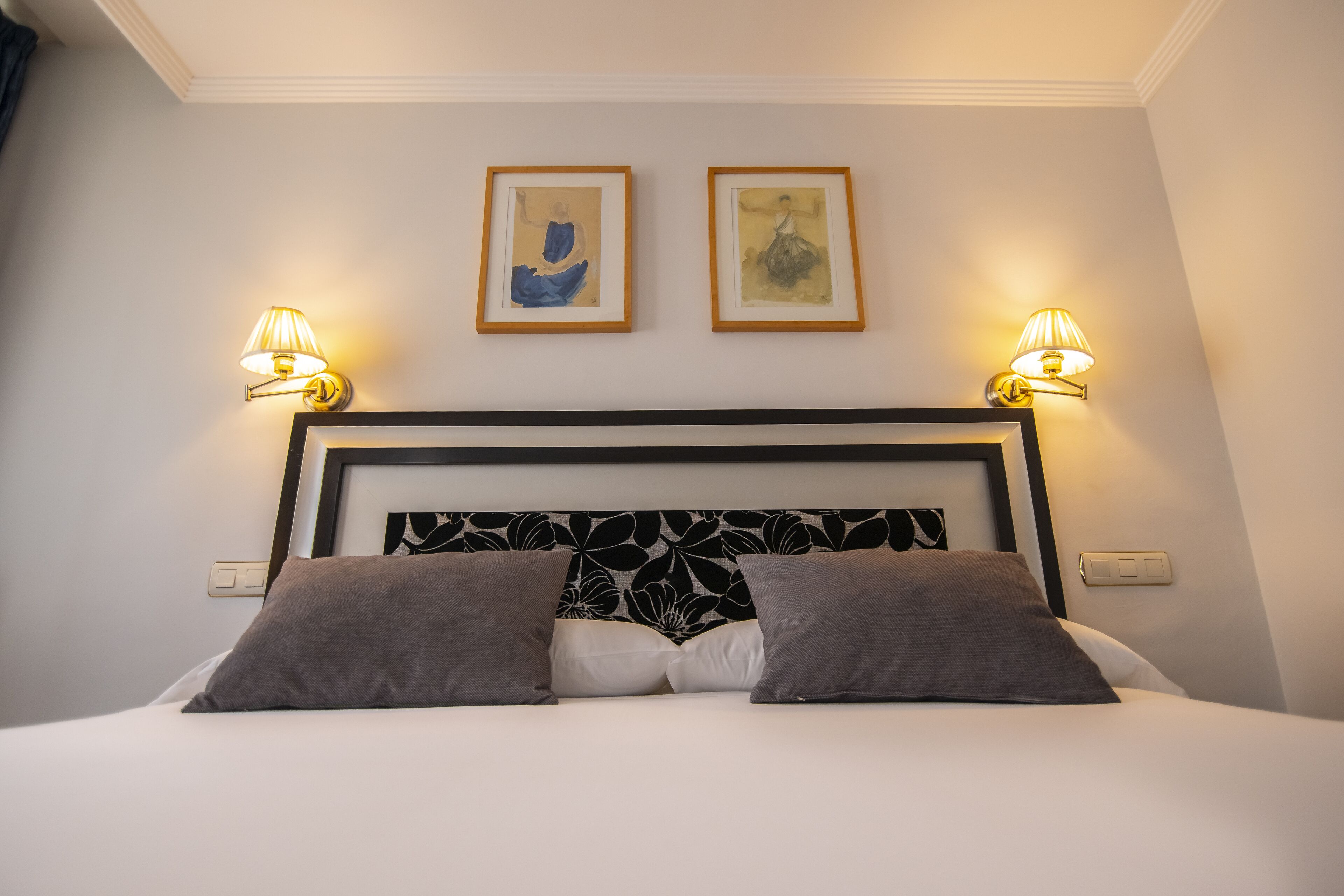 A neatly made bed with a black and white headboard, two framed artworks on the wall, and wall-mounted lamps.