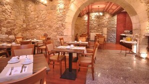 Restaurante com paredes de pedra e mesas de madeira. As mesas estão montadas com pratos, talheres e guardanapos. Há uma área de buffet com balcão e diversos alimentos.