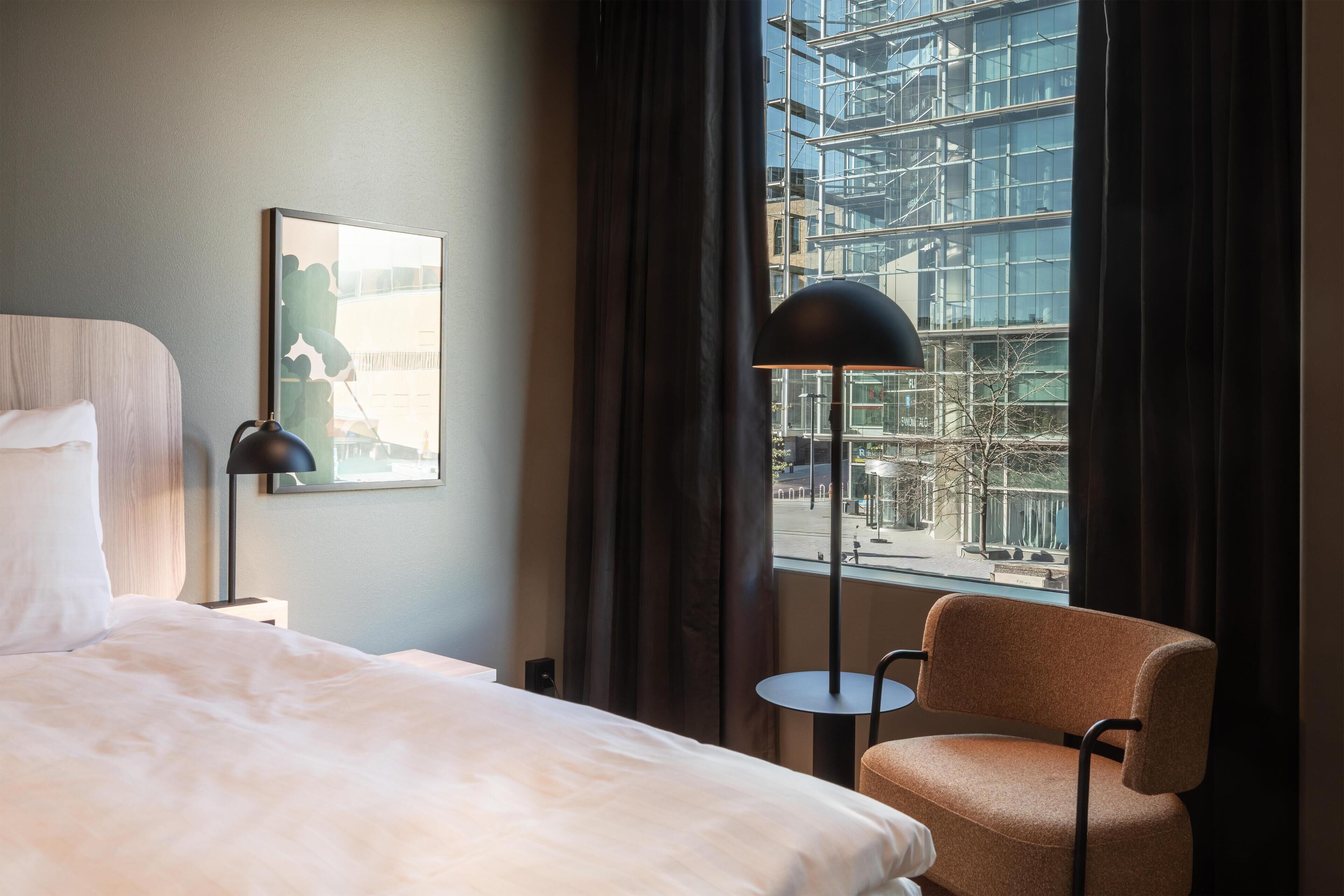 A hotel room with a bed, a chair, a lamp, and a large window with a view of a building under construction.
