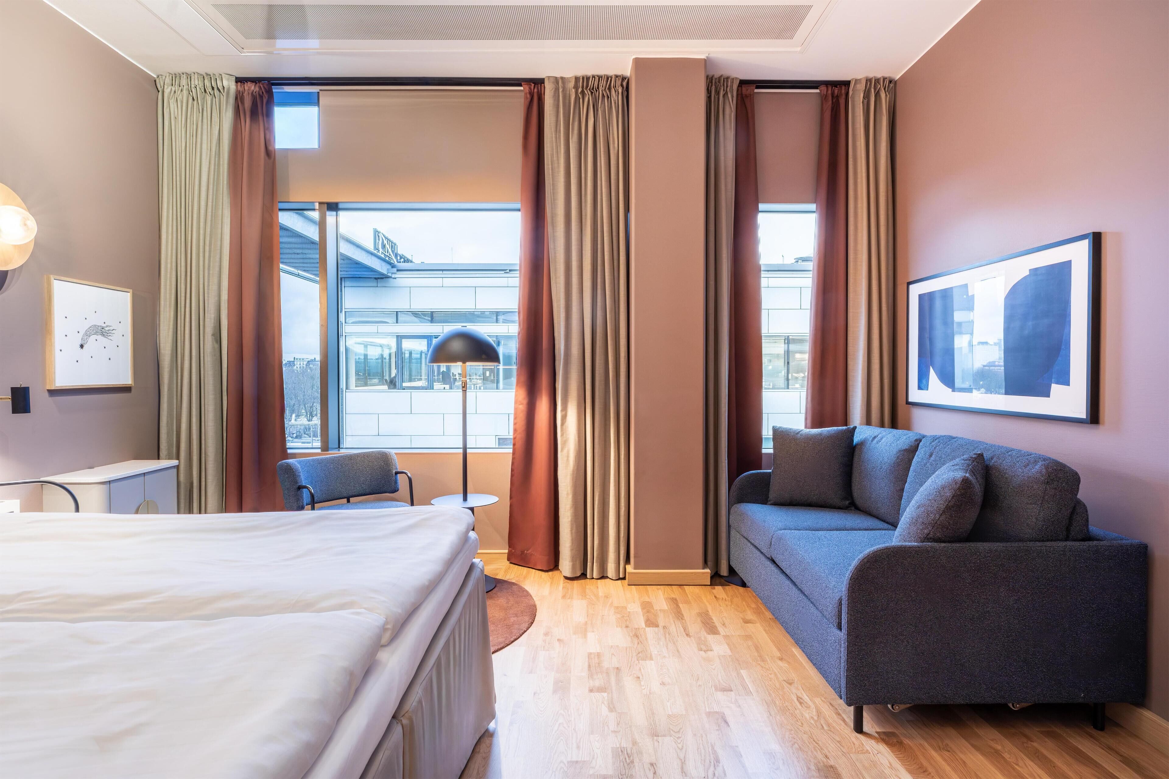 Ett hotellrum med en säng, en soffa, ett skrivbord och ett stort fönster med gardiner.