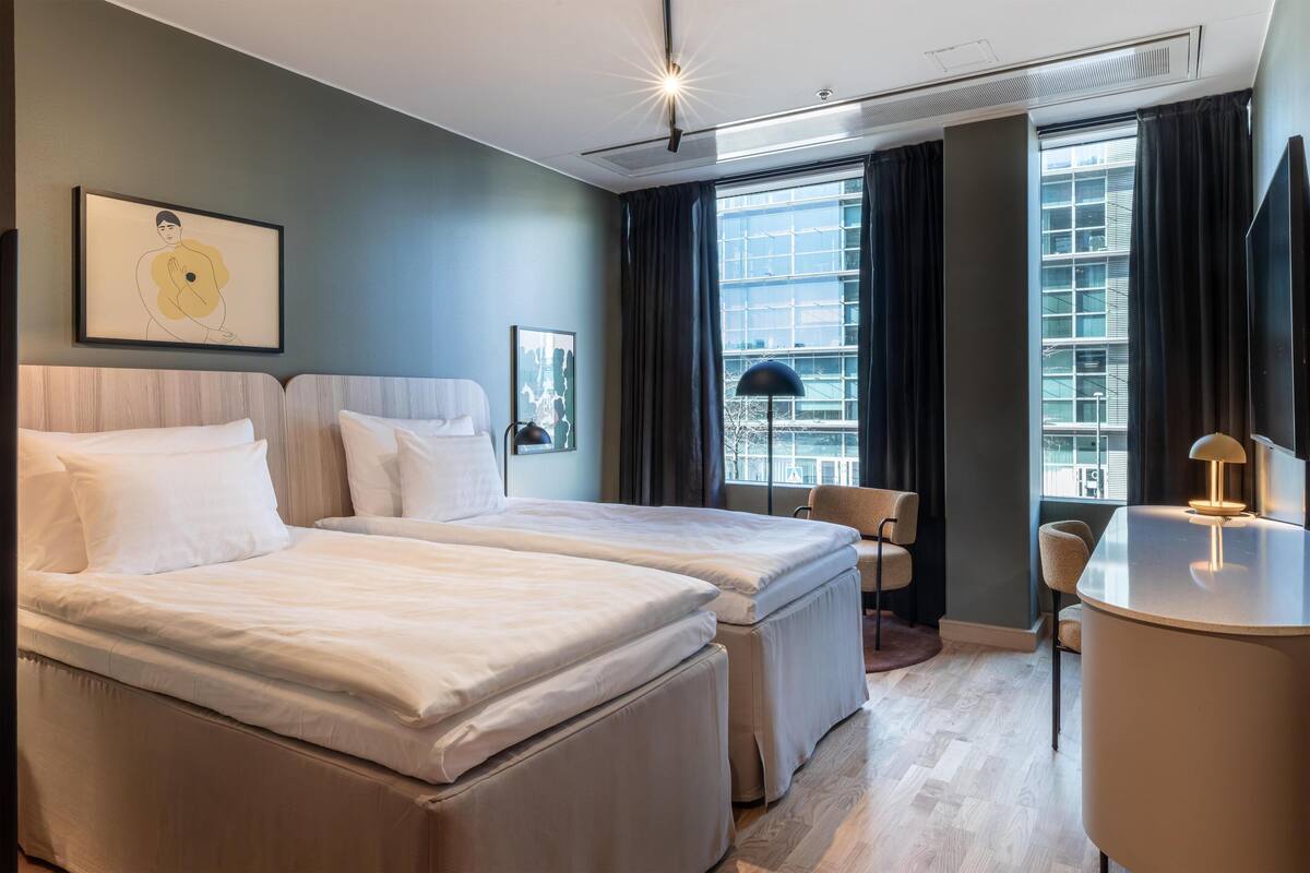 A hotel room with two beds, a desk, a chair, and large windows with curtains.