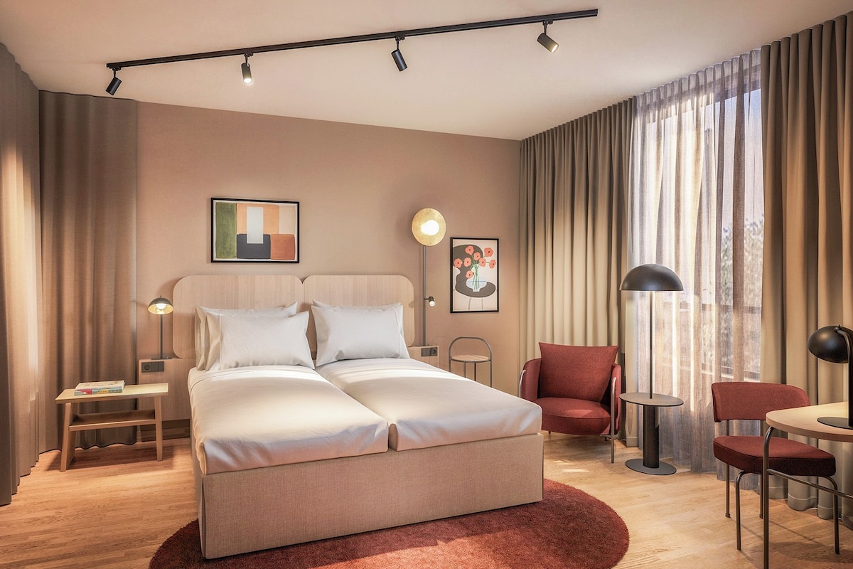 A modern hotel room with a bed, two bedside tables, a chair, a desk, and a large window with curtains.