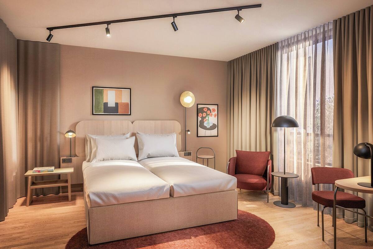 A modern hotel room with a bed, two bedside tables, a chair, a desk, and a large window with curtains.