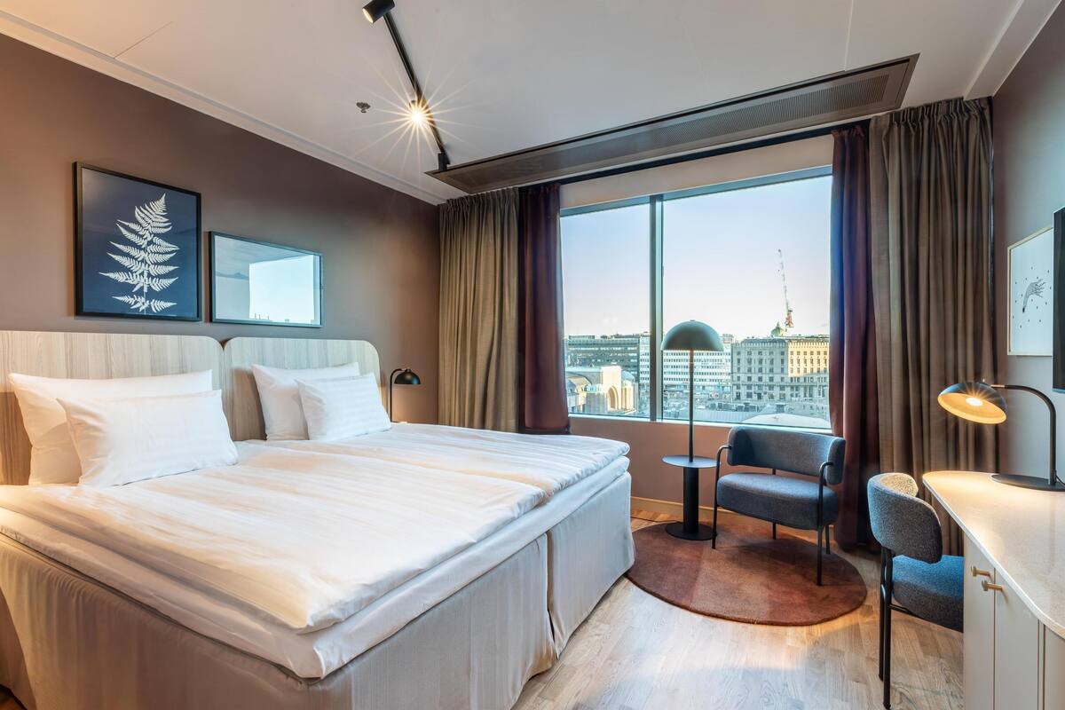 A hotel room with a large bed, a desk with a chair, a view of a cityscape through large windows, and a ceiling light.