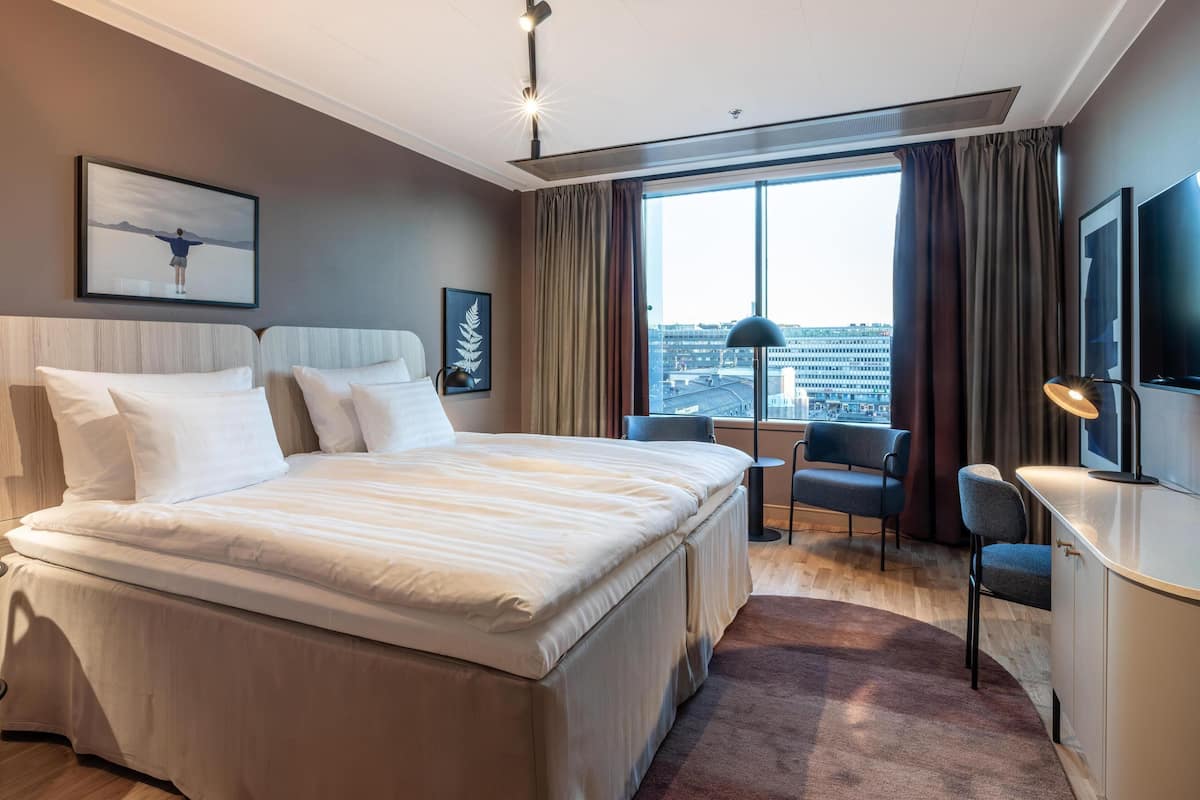 A modern hotel room with a large bed, a desk, and a chair, a large window with curtains, and a city view.