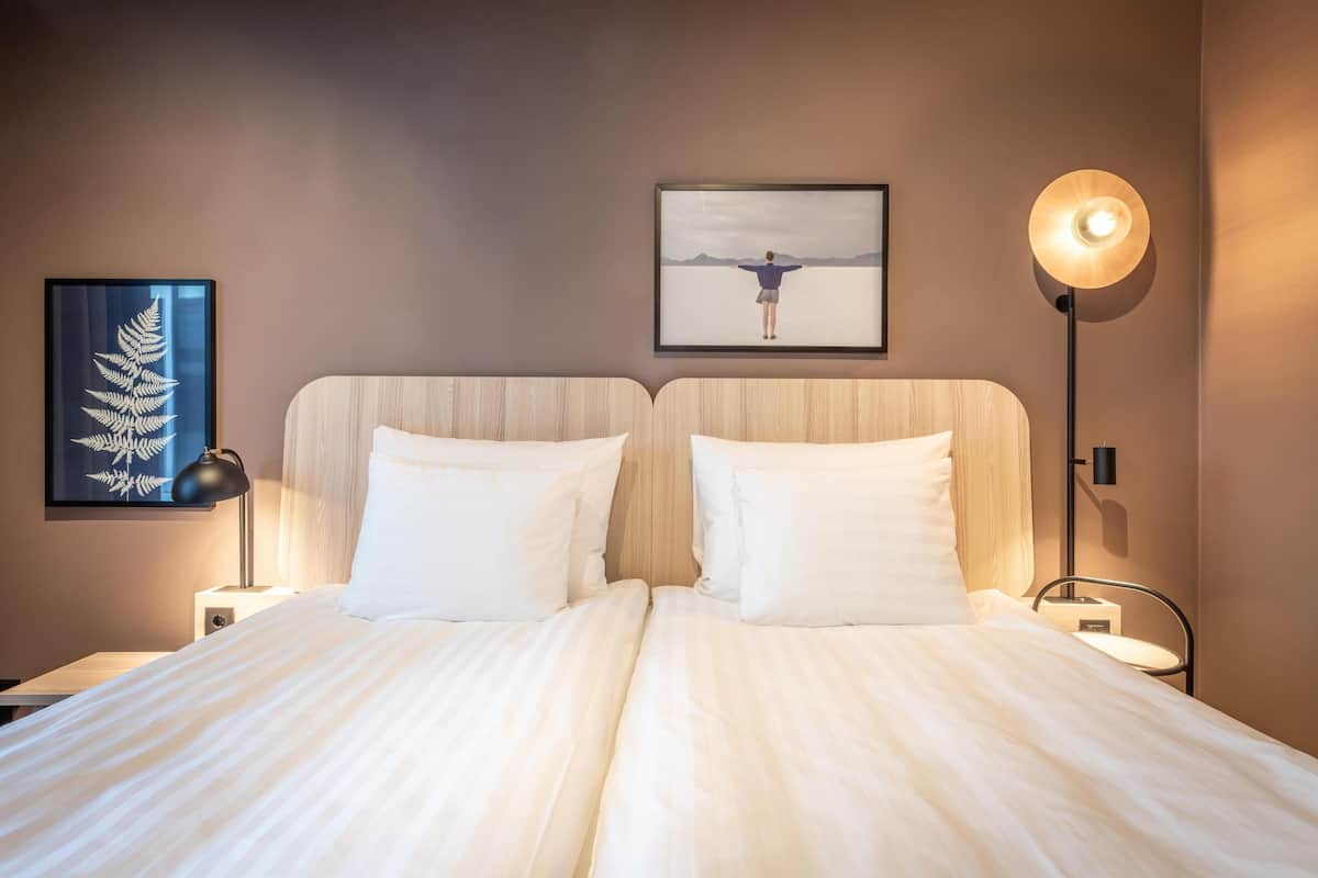A double bed with white linens, two bedside tables, a framed artwork of a person with arms outstretched, a floor lamp, and a wall-mounted picture of a fern.