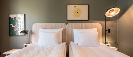 A hotel room with two beds, a framed artwork on the wall, a desk with a lamp, and a window with a view of buildings.