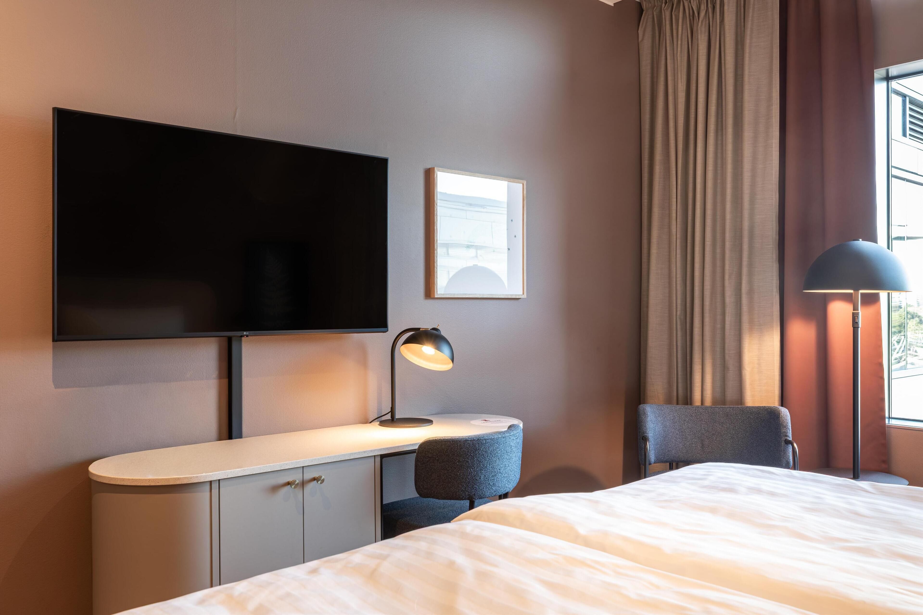 A hotel room with a bed, a desk with a lamp, a TV, and a window with curtains.