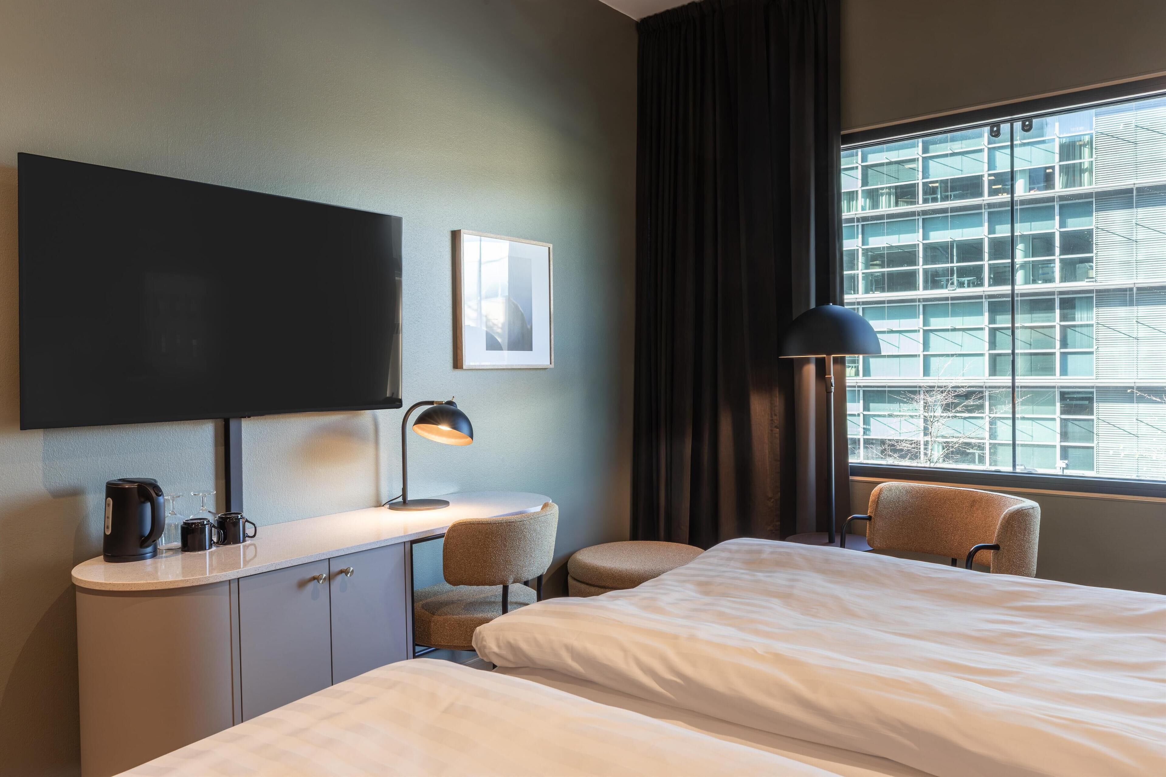 Habitación de hotel con cama, escritorio con lámpara, cafetera y televisión en la pared.