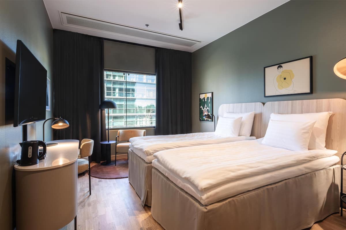 A hotel room with two beds, a desk with a kettle, a chair, a desk lamp, and a large window with curtains.