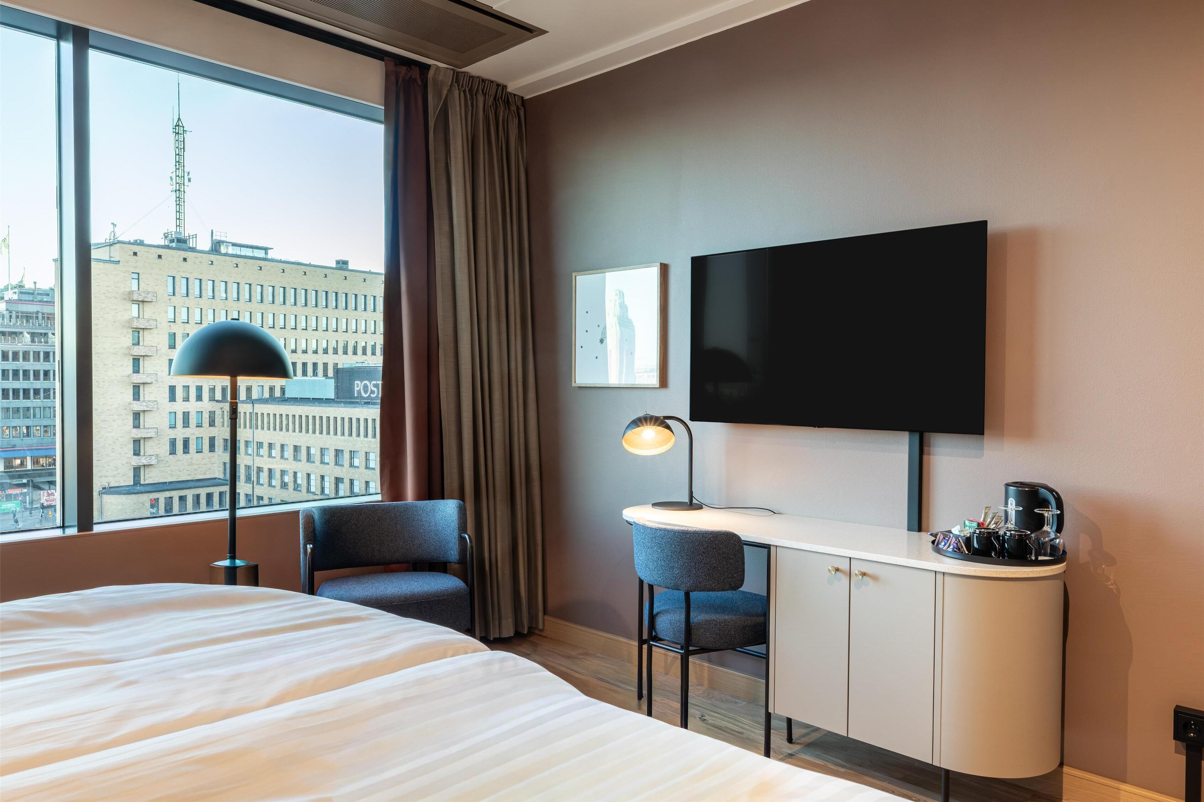 Ett hotellrum med en säng, ett skrivbord, stolar, en tv och utsikt över staden.