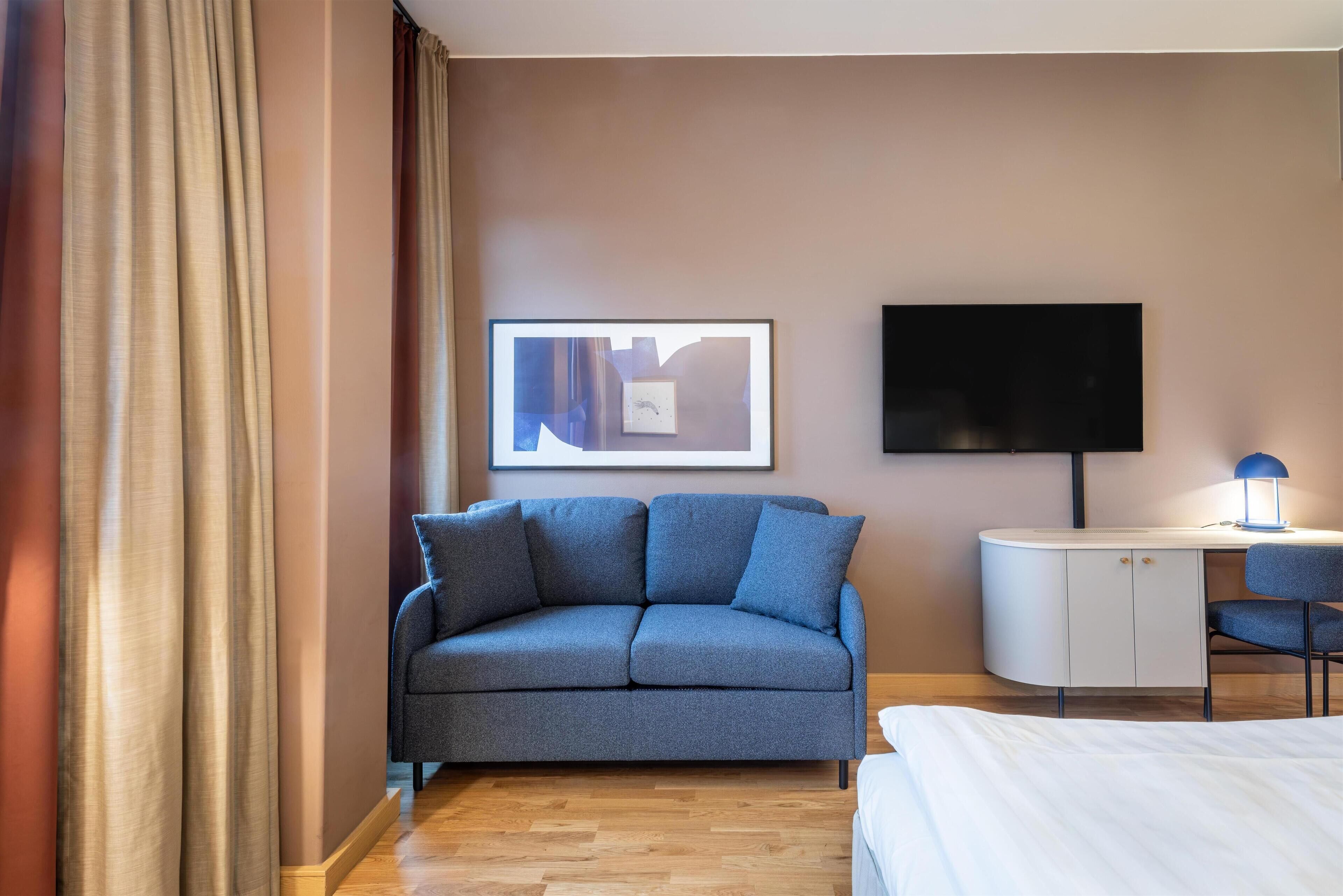 Een moderne hotelkamer met een grijze bank, een wit bureau met een lamp, een flatscreen-televisie en een ingelijst kunstwerk aan de muur.
