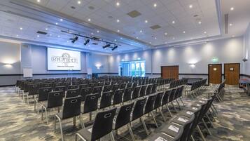 A conference room with rows of chairs, a projector screen displaying 'Grand Magic Hotel', and a carpeted floor.