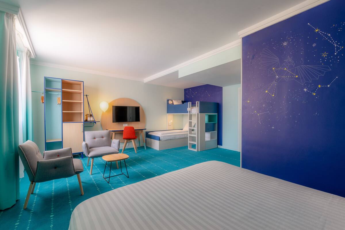 A modern hotel room with a bed, a desk, a chair, a TV, and a wardrobe.