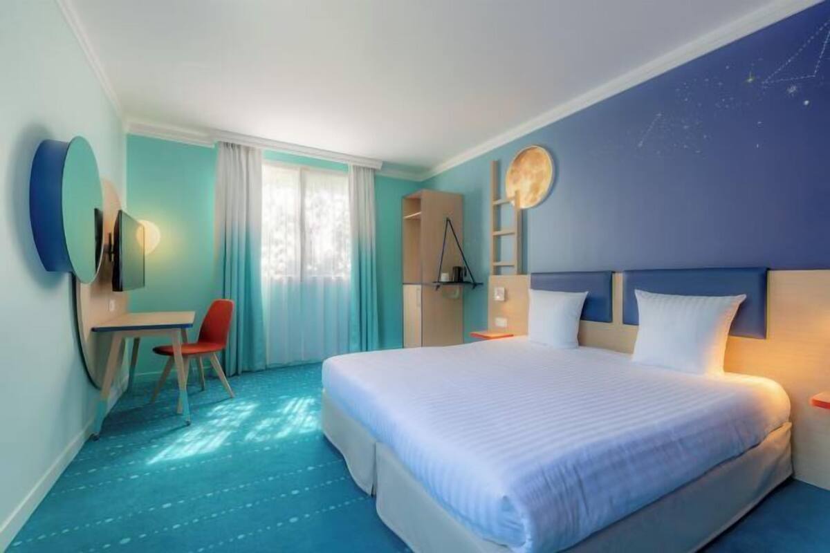 A hotel room with a large bed, a desk, and a chair. There is a window with curtains, a wardrobe, and a wall-mounted TV.
