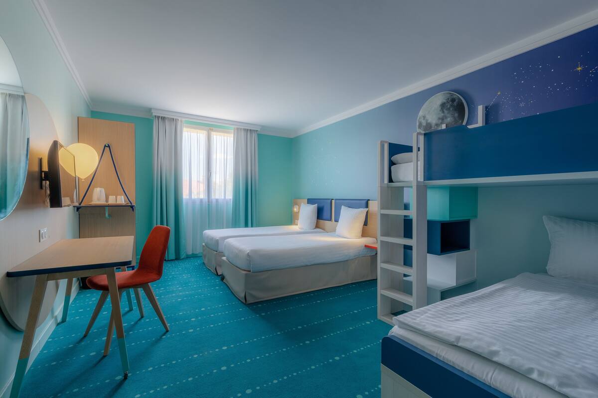 A hotel room with two beds, a desk, a chair, and a bunk bed.