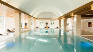 Una piscina interior con agua cristalina, ambiente iluminado por el sol y zonas de descanso con plantas en macetas.
