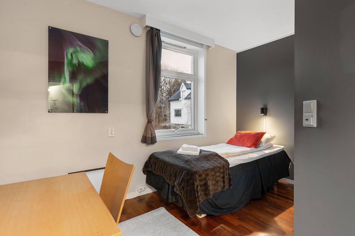 Un dormitorio con cama, ventana, una silla, una mesa y un cuadro en la pared.