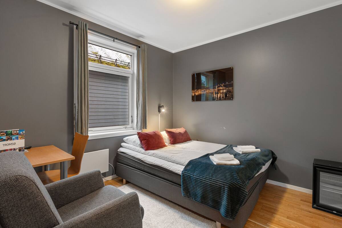 Un dormitorio con cama, una silla, una mesa, una ventana con cortinas, un radiador y una obra de arte colgada en la pared.
