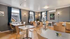 Un comedor moderno con varias mesas y sillas, ventanas grandes con cortinas y unidades de aire acondicionado fijadas a la pared.