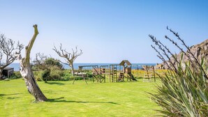 Un espacio cubierto de hierba con una estructura de madera, un árbol inclinado y vistas al mar.