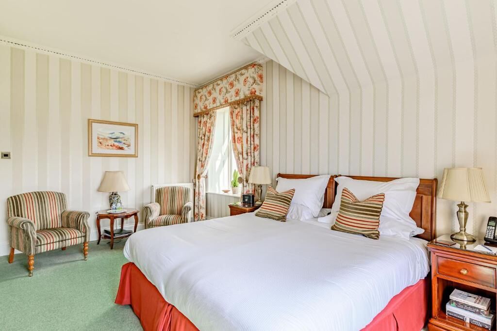 Quarto de hotel com uma cama grande, duas poltronas, uma mesa pequena e uma mesinha de cabeceira com abajur.