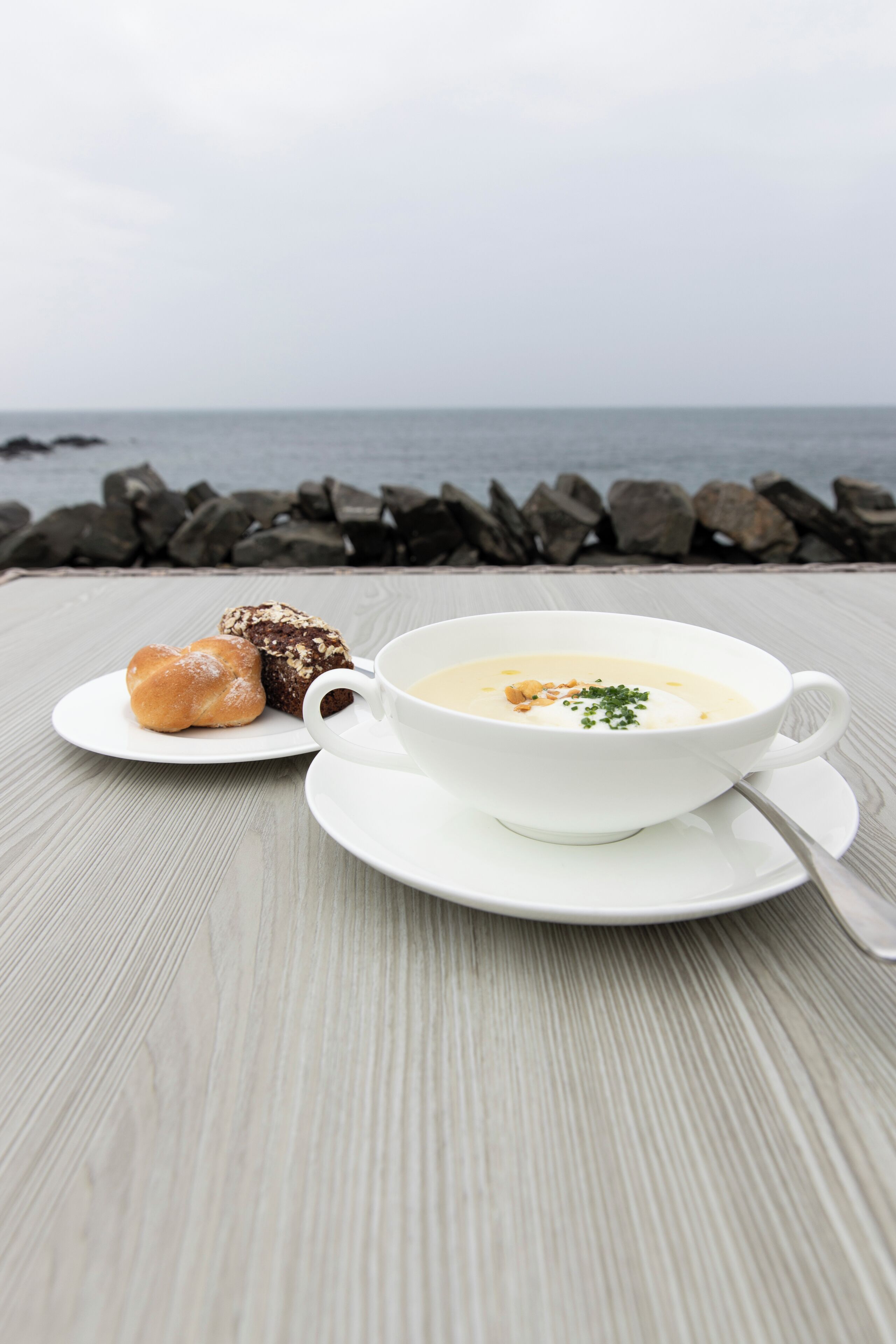 A table with a bowl of soup and two pastries on a wooden table by the sea.