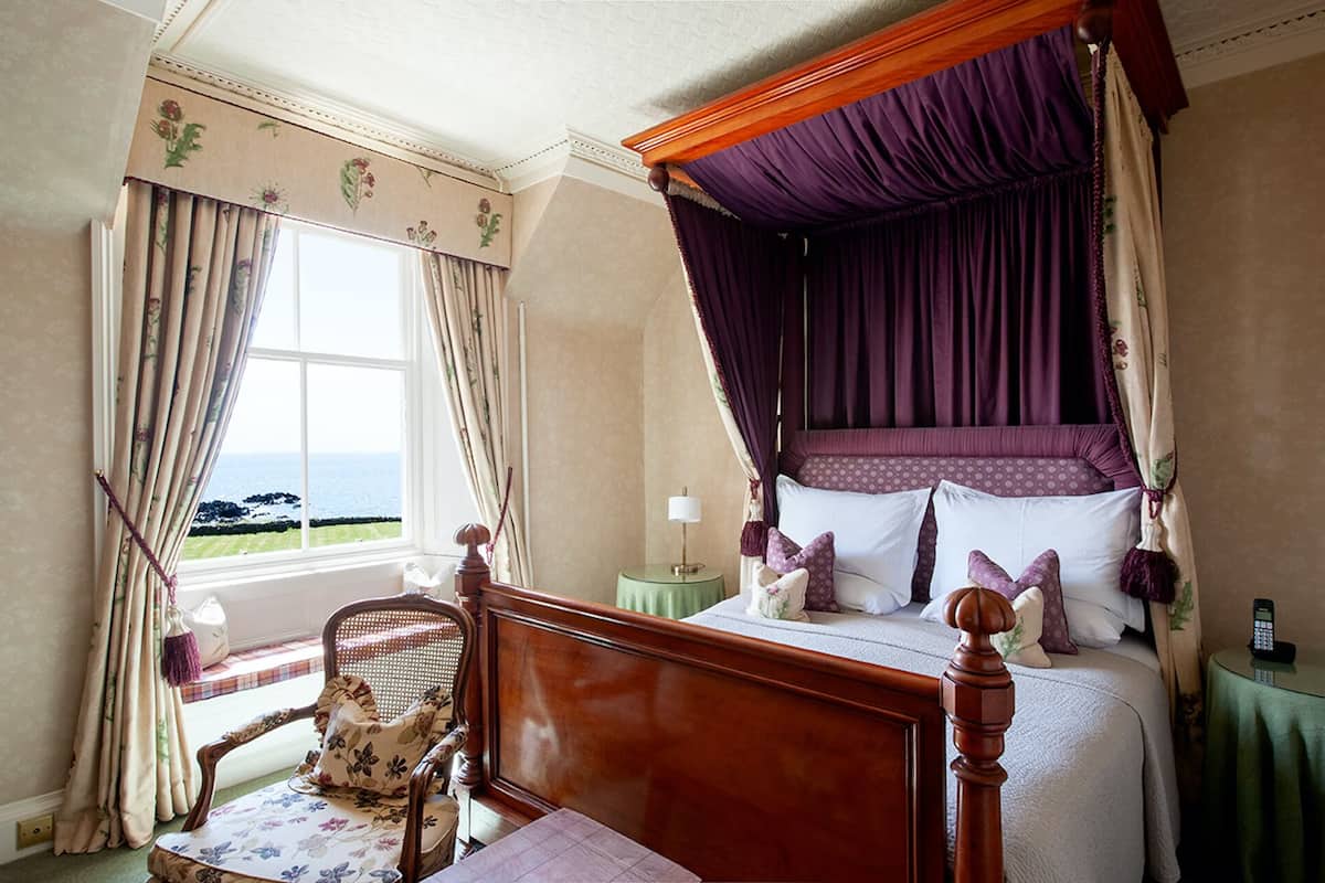 A bedroom with a four-poster bed, a chair, a bedside table, and a view of the sea through a window.