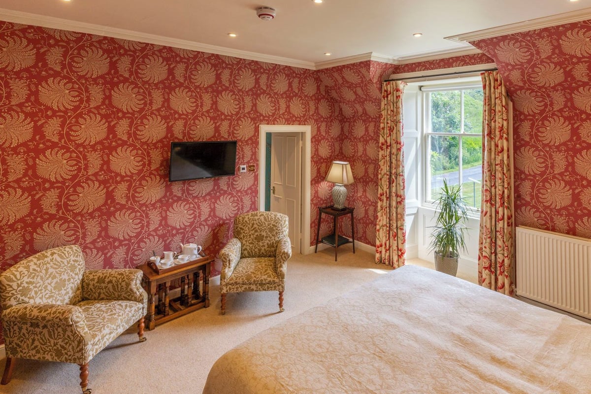 A bedroom with a large bed, two armchairs, a small table with tea set, a TV mounted on the wall, and a window with curtains.