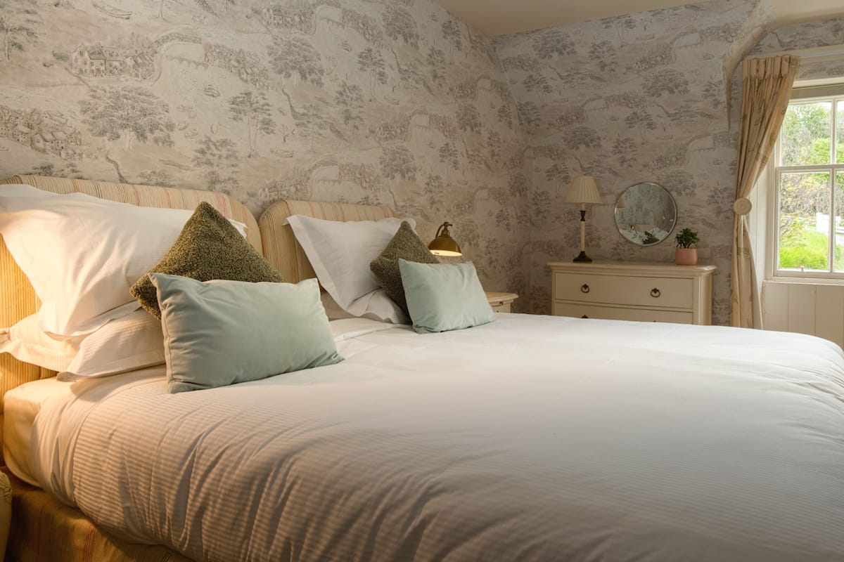 A bedroom with a large bed, a nightstand, a lamp, a mirror, and a window with curtains.