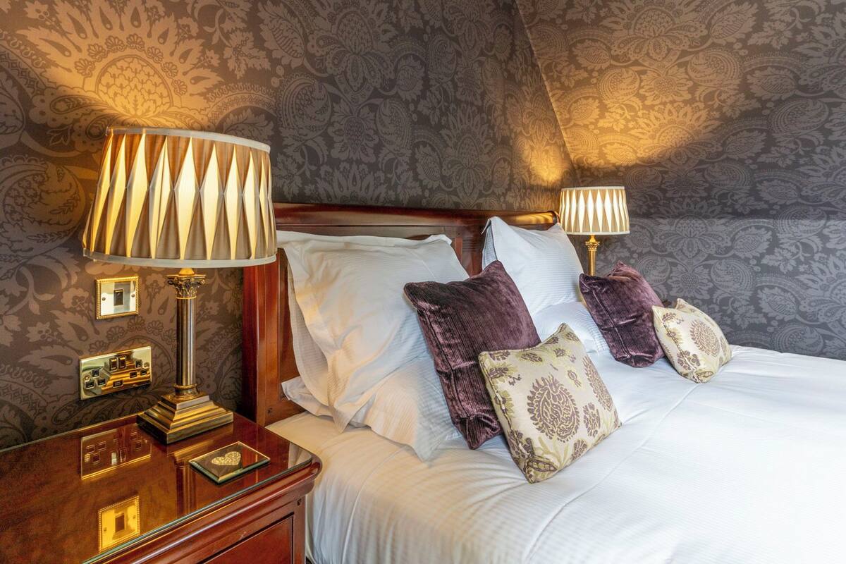 A neatly made bed with white linens and multiple pillows, including a decorative one. There are two bedside lamps with lampshades, a wooden nightstand with a mirror and a small table, and a wall with a patterned wallpaper.