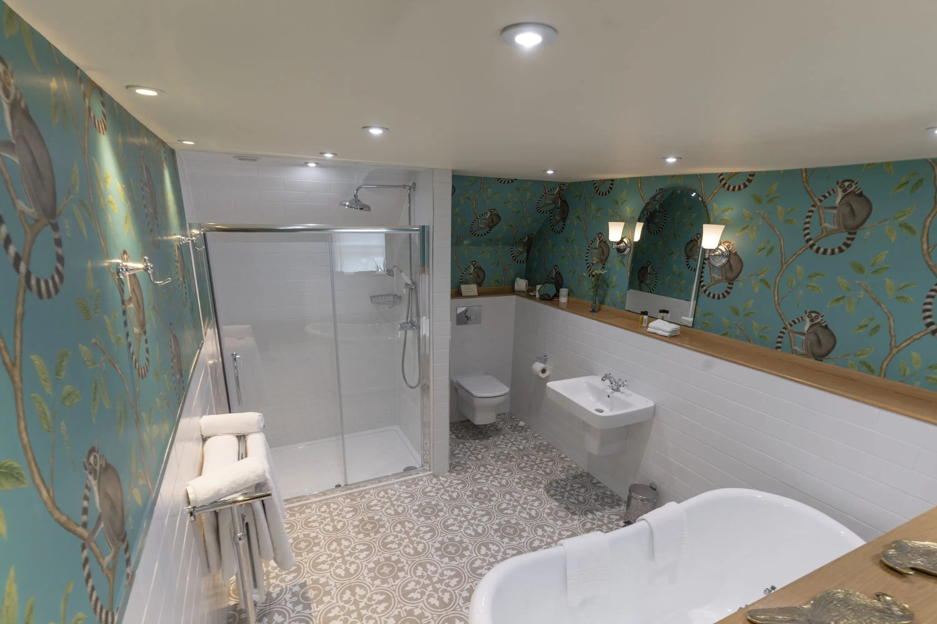 Un baño con ducha de vidrio, una bañera blanca, un lavamanos y un inodoro. Las paredes tienen papel tapiz con motivos de animales.