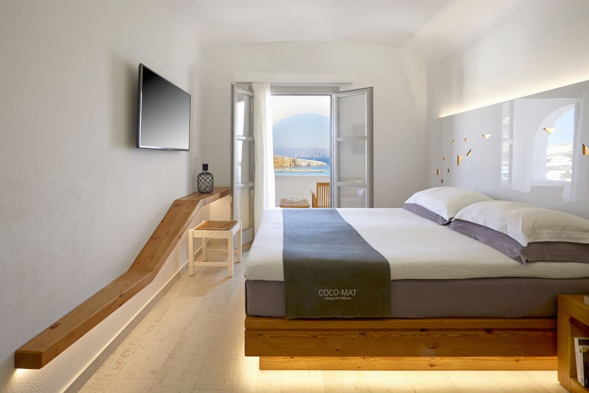 Un dormitorio moderno con cama, televisor montado en la pared y vista al mar a través de una puerta abierta.