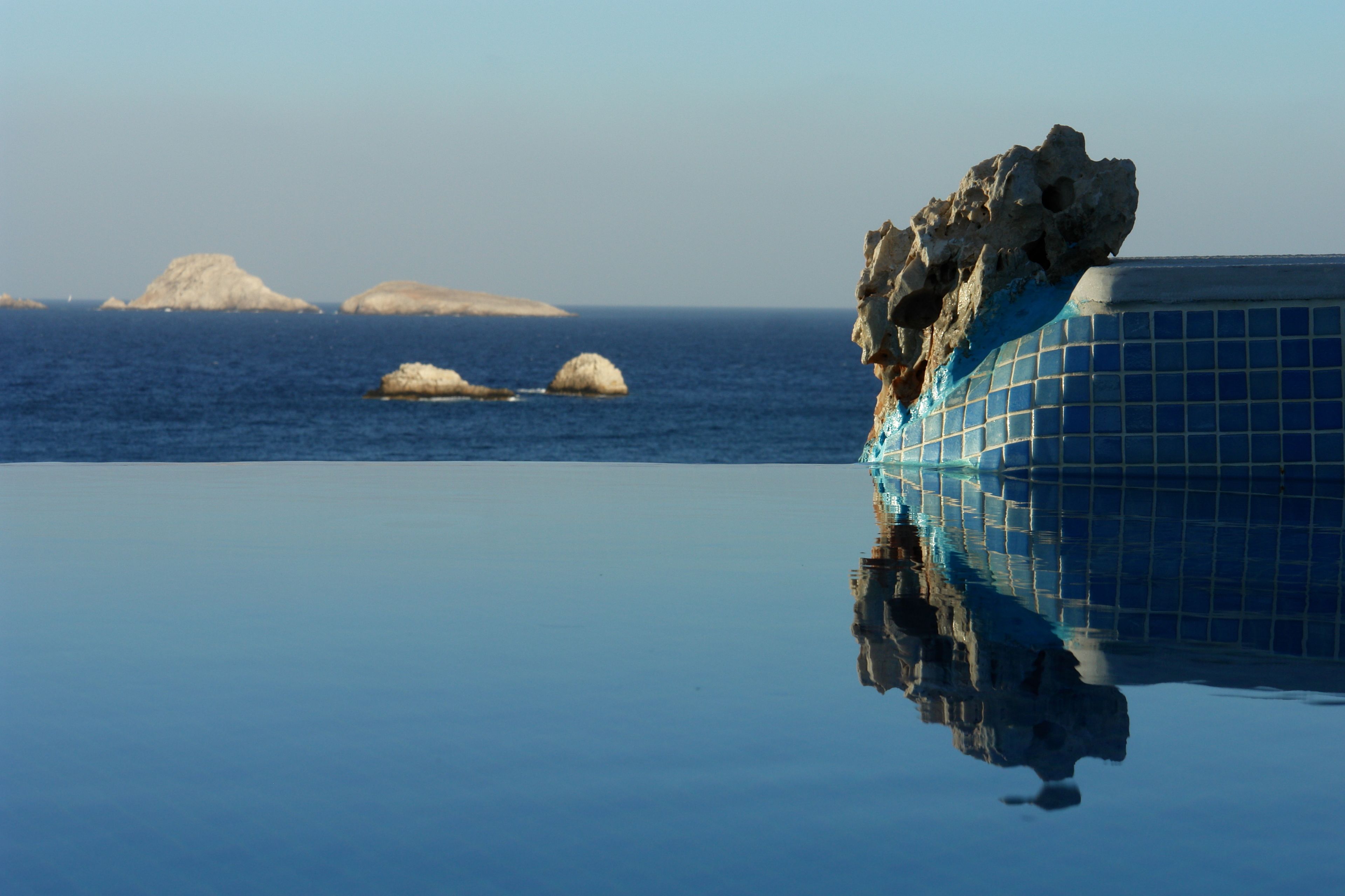 A clear view of a swimming pool with a rock formation on the right and distant islands in the background.