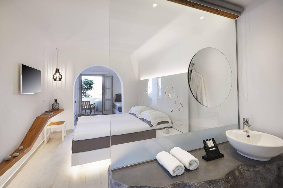 Habitación de hotel moderna con una cama grande, un banco de madera, un televisor, un espejo grande, un espejo redondo, un lavamanos y vistas al exterior.