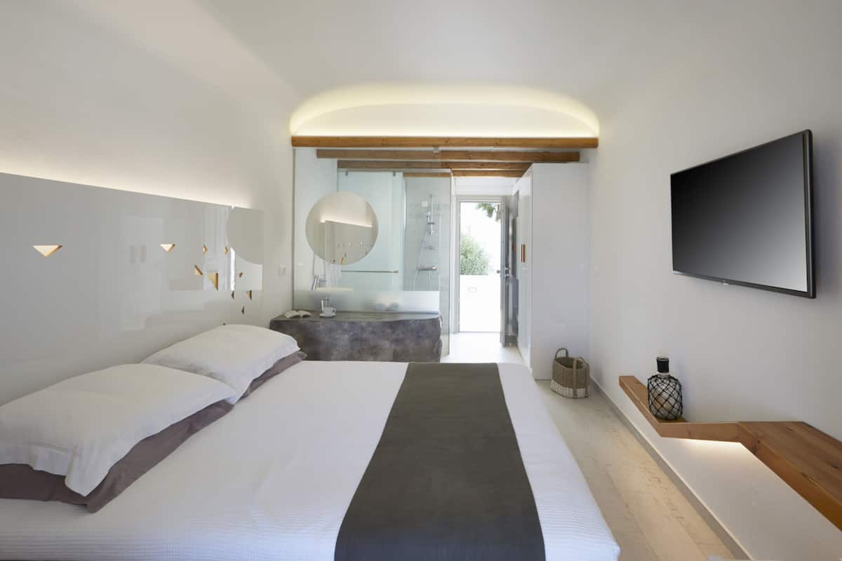 Un dormitorio moderno con una cama grande, un televisor de pantalla plana, un cabecero de madera, una mesita de noche y un baño visible a través de una puerta abierta.
