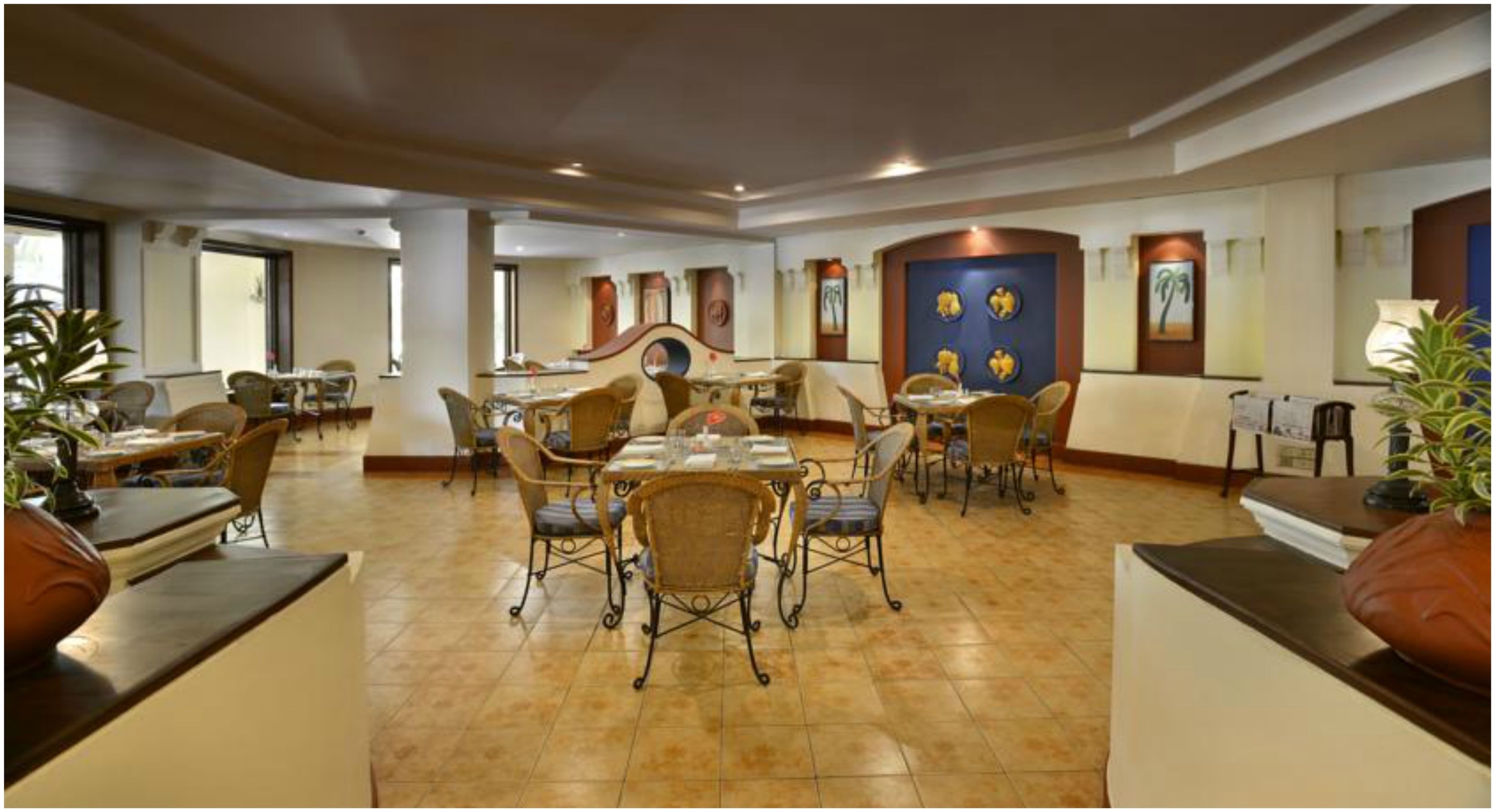 A hotel dining area with multiple tables and chairs, a central decorative wall, and potted plants.